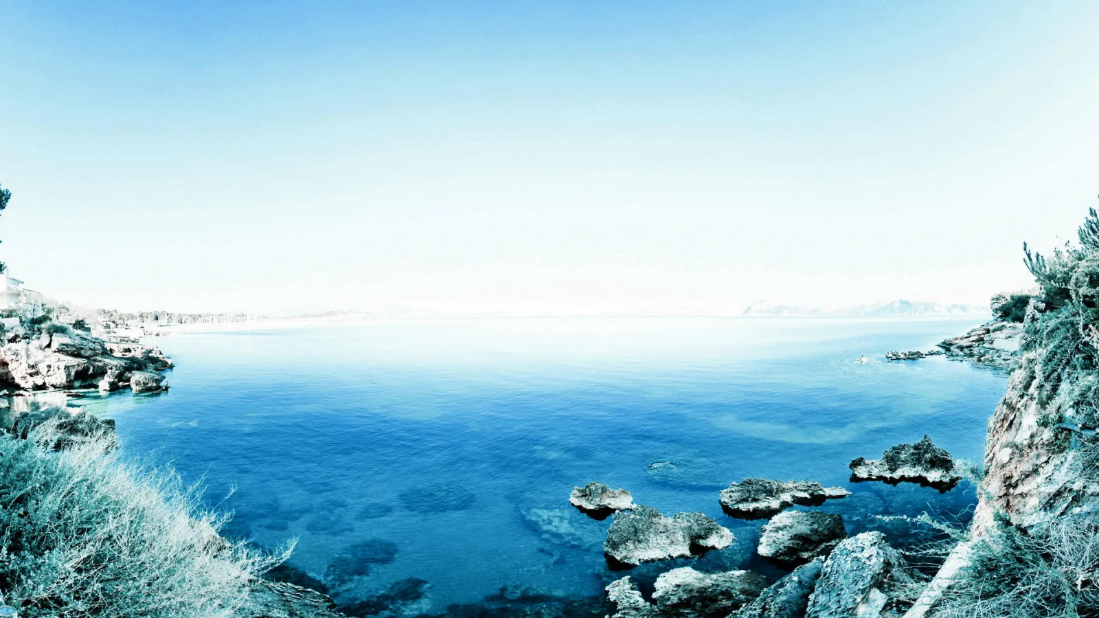 Ruhige blaue Meeresbucht mit Felsen im Vordergrund und hellem Himmel darüber.
