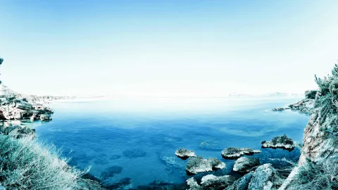 Ruhige blaue Meeresbucht mit Felsen im Vordergrund und hellem Himmel darüber.