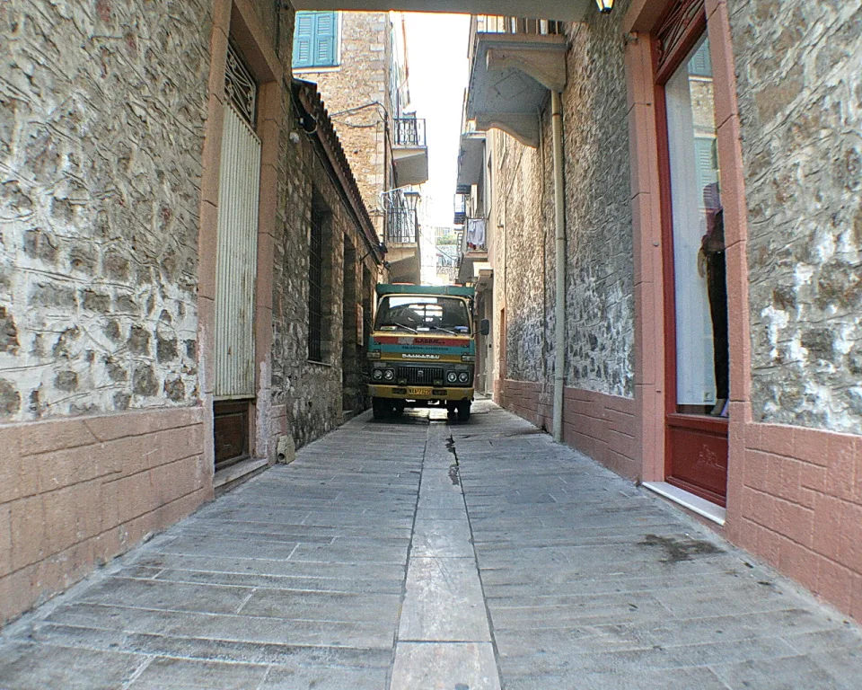 Schmale Gasse mit Steinmauern und rötlichen Sockeln, am Ende steht ein kleiner grün-gelber LKW zwischen den Hauswänden.