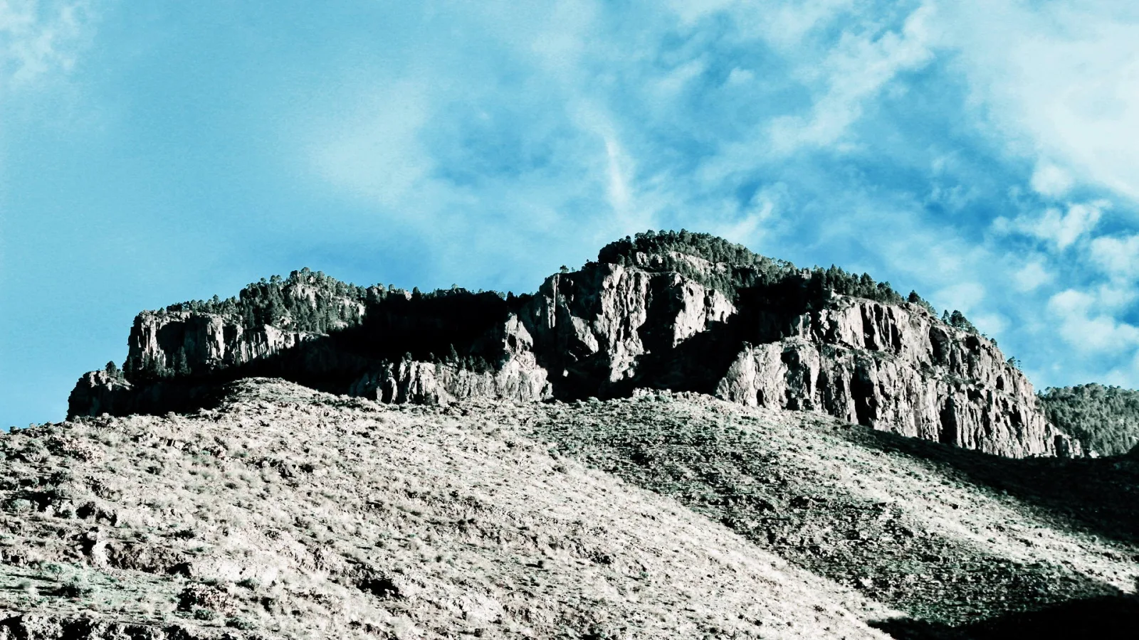 Steiler Felsrücken mit lichtem Kiefernbewuchs ragt über einen trockenen, hellbraunen Hang unter einem blauen Himmel mit Schleierwolken.