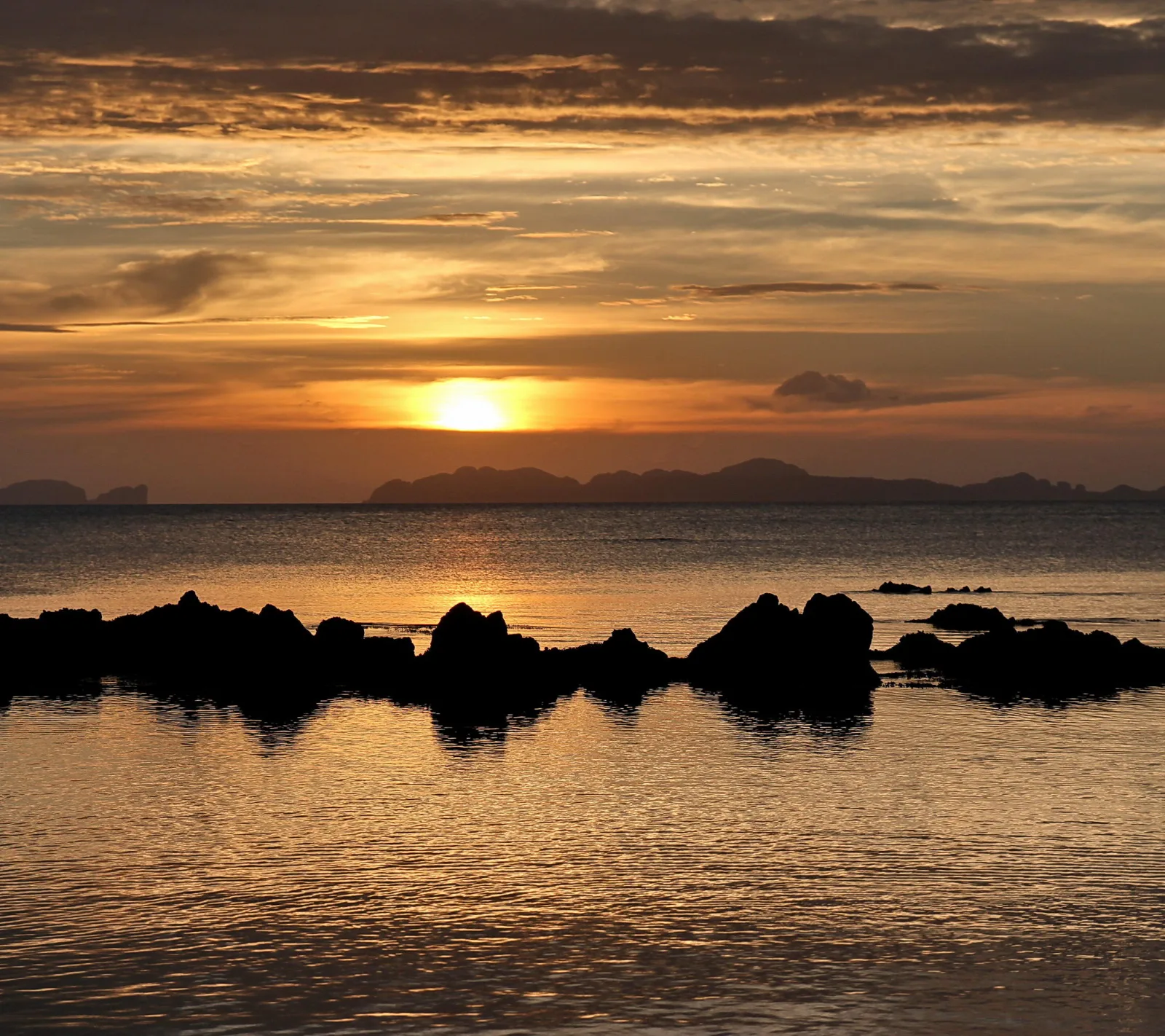 Die Sonne steht tief über dem Meer und hinter Inseln am Horizont. Dunkle Felsen zeichnen sich als Silhouetten im spiegelnden Wasser ab, der Himmel leuchtet orange und grau.