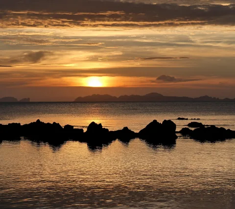 Die Sonne steht tief über dem Meer und hinter Inseln am Horizont. Dunkle Felsen zeichnen sich als Silhouetten im spiegelnden Wasser ab, der Himmel leuchtet orange und grau.