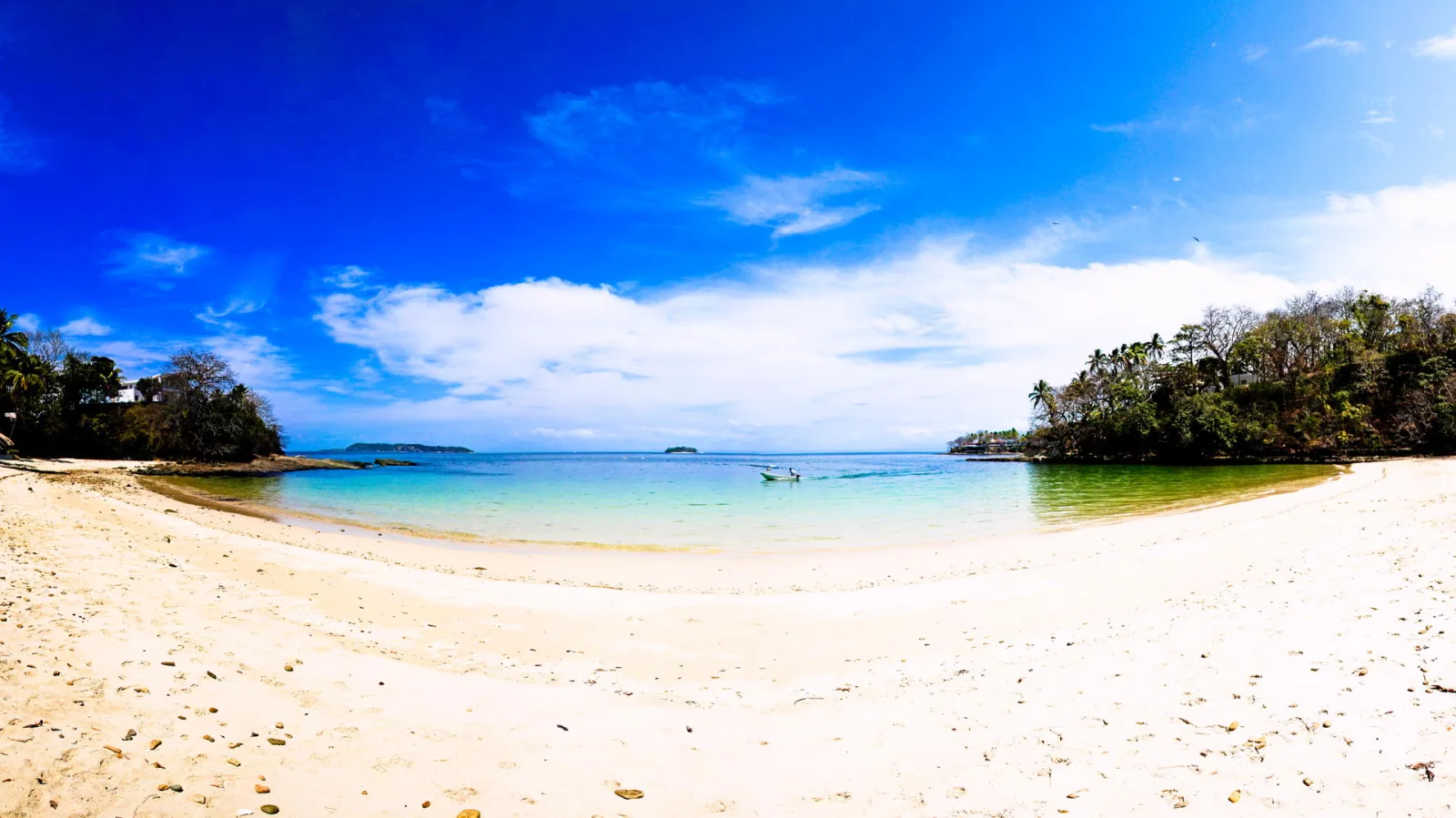 Weiter Sandstrand im Vordergrund, davor eine flache türkisfarbene Bucht mit einem kleinen Boot, links und rechts bewachsene Küstenabschnitte unter blauem Himmel mit Wolken.