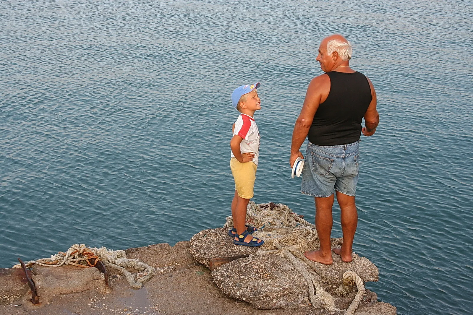 Ein älterer Mann mit weißem Haar, barfuß und in Jeansshorts, steht mit einem Jungen in blauer Basecap auf einer Steinmole am Meer, die beiden schauen sich an.