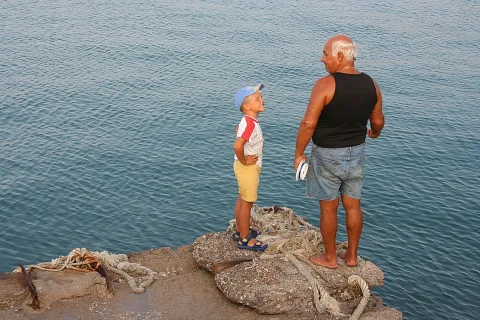 Ein älterer Mann mit weißem Haar, barfuß und in Jeansshorts, steht mit einem Jungen in blauer Basecap auf einer Steinmole am Meer, die beiden schauen sich an.