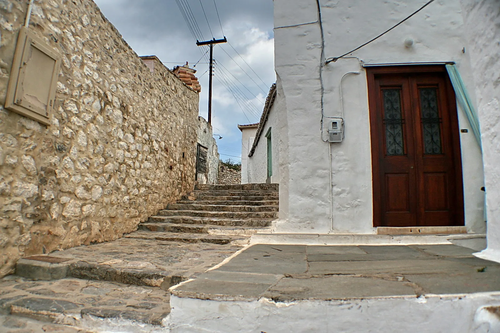 Schmale Gasse in einem mediterranen Dorf: links eine grobe Bruchsteinmauer, rechts eine weiß getünchte Hauswand mit einer dunkelbraunen Doppeltür, davor eine Treppe und ein Plattenweg, darüber bewölkter Himmel und ein Strommast.