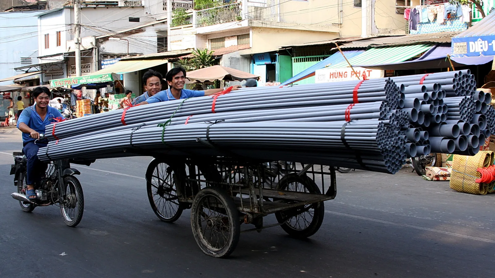 Zwei Männer auf einem Motorrad und einem Handkarren mit einem langen Stapel grauer Kunststoffrohre, die mit roten Gurten gesichert sind, auf einer Straße in Ho-Chi-Minh-Stadt. Im Hintergrund Wohnhäuser, Markisen und Verkaufsstände.