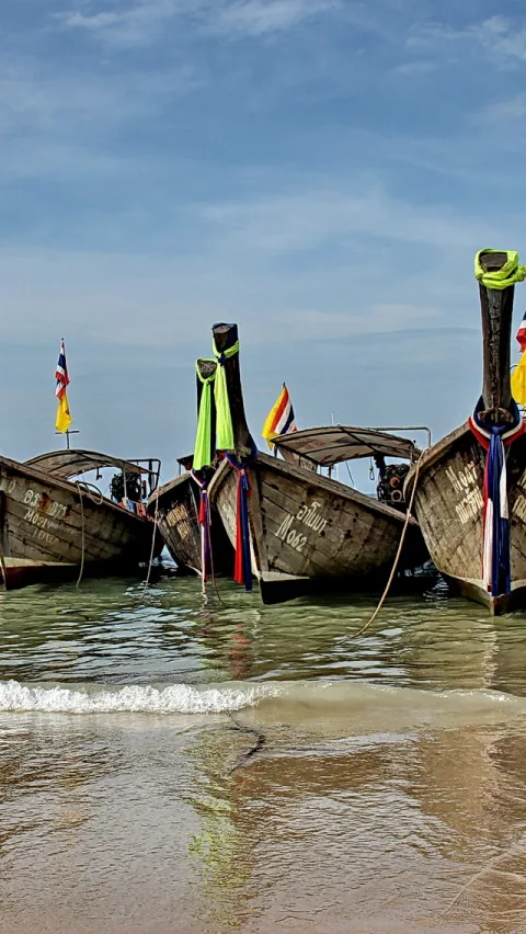 Mehrere hölzerne Longtailboote liegen im flachen Wasser vor einem Sandstrand, mit bunten Bändern am Bug und einer thailändischen Flagge links, darüber blauer Himmel mit Wolken.