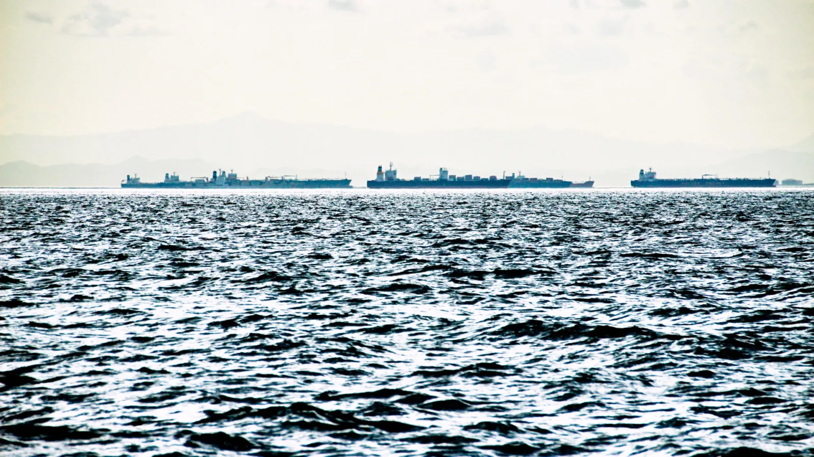 Drei große Frachter und Tanker liegen in weiter Entfernung auf dem Meer vor einer Küste mit blassen Bergen am Horizont, im Vordergrund bewegte dunkle Wellen unter hellem, diesigem Himmel.
