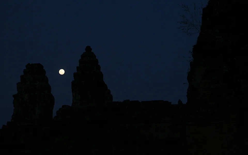 Dunkle Silhouetten mehrerer Tempeltürme vor einem nachtblauen Himmel, links neben den Türmen steht der helle Vollmond.