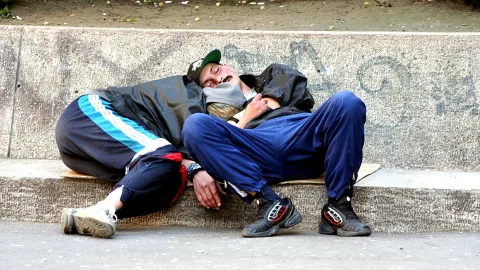 Zwei Männer in Jacken und Jogginghosen liegen auf einem betonierten Randstein und schlafen, vor einer grauen Mauer mit Graffiti.