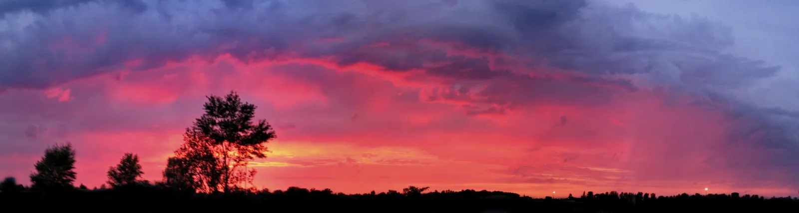 Panoramaaufnahme eines Sonnenuntergangs mit rot-pink leuchtenden Wolken und dunklen Baumsilhouetten am Horizont.