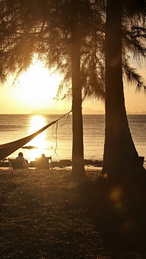 Hängematte zwischen Kiefernstämmen an einem Strand, im Hintergrund die untergehende Sonne über dem Meer, zwei Personen sitzen auf Liegestühlen.