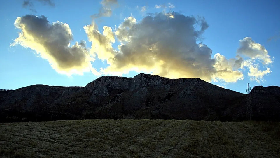 Felsiger Bergkamm vor blauem Himmel mit angestrahlten Wolken, davor eine flache Grasebene