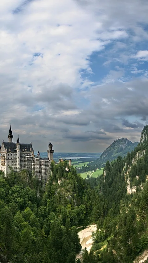 Schloss Neuschwanstein mit hellen Türmen und Baugerüsten auf einem bewaldeten Felsen, darunter ein Tal mit Fluss und Wiesen, darüber bewölkter Himmel.