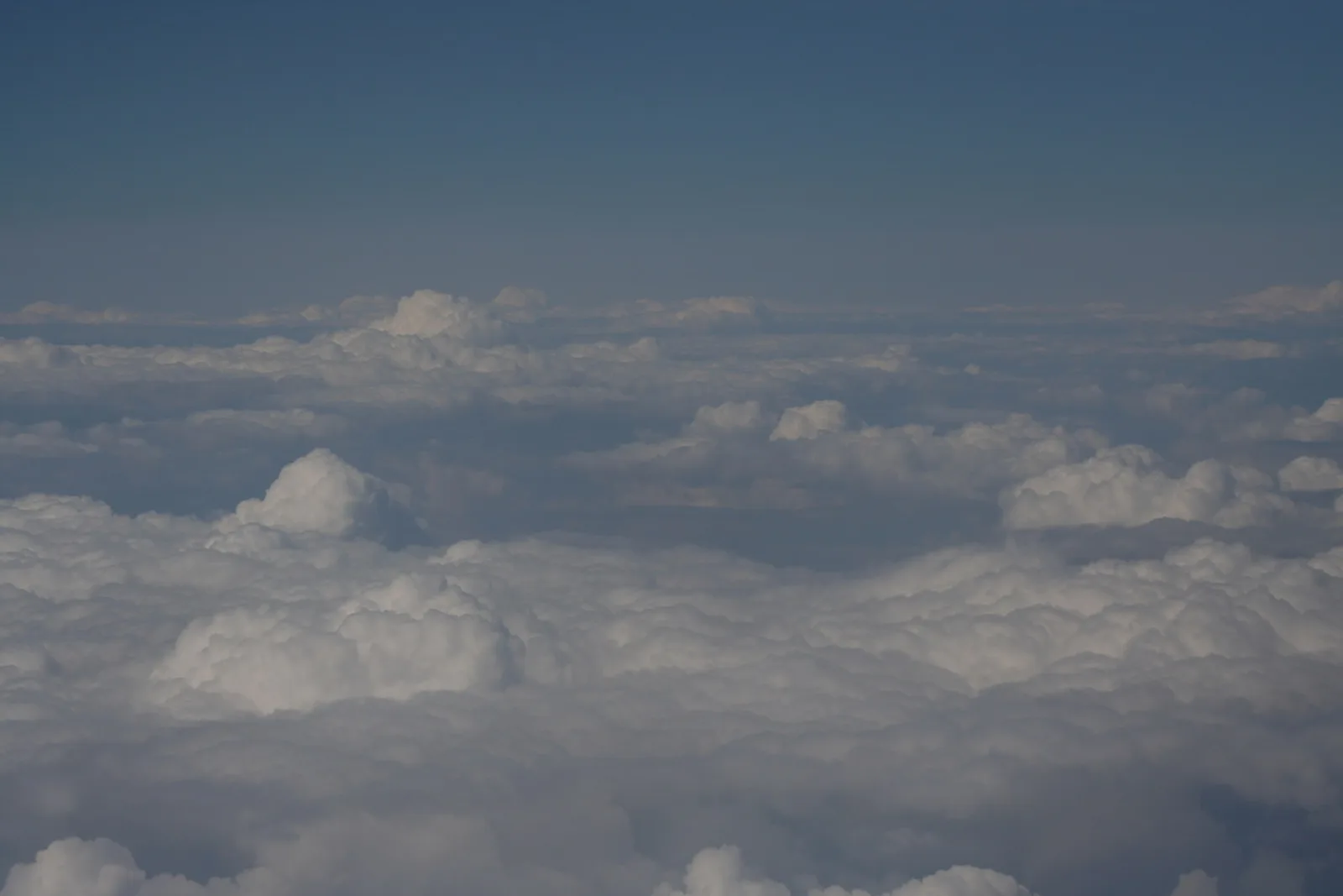Luftaufnahme von oben auf eine weiße Wolkendecke mit aufgetürmten Kumuluswolken, darüber ein blassblauer Himmel.