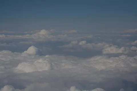 Luftaufnahme von oben auf eine weiße Wolkendecke mit aufgetürmten Kumuluswolken, darüber ein blassblauer Himmel.