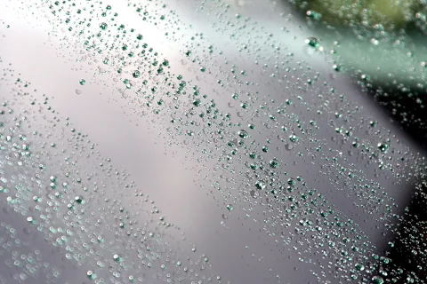 Viele kleine Wassertropfen perlen auf einer grauen, glatten Glasoberfläche, durchsetzt mit grünlichen Lichtreflexen.