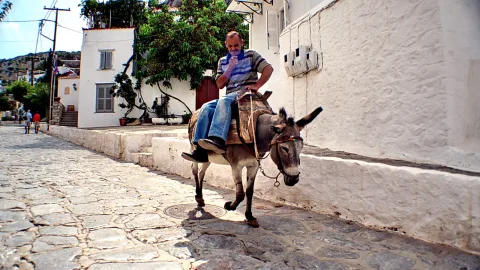 Ein älterer Mann mit Schnurrbart und gestreiftem Hemd reitet auf einem grauen Esel über eine gepflasterte Gasse, an weiß getünchten Häusern vorbei.