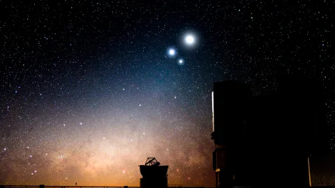 Nachthimmel voller Sterne über der Milchstraße, rechts zwei dunkle Gebäude eines Observatoriums, unten links ein Teleskop mit Schüssel, am Horizont ein warmer orangefarbener Schimmer.