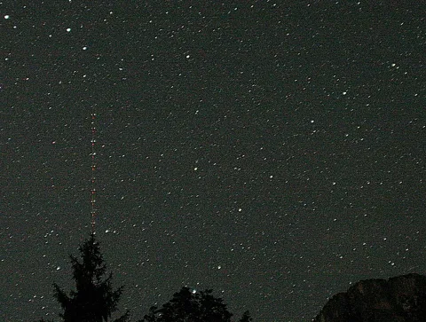 Nachtaufnahme eines dicht besternten Himmels. Links ragt ein dünner Funkmast mit roten Warnlichtern auf, unten sind Nadelbaumsilhouetten und rechts ein dunkler Felsrücken zu sehen.