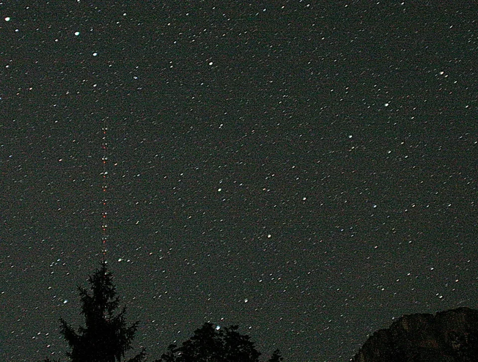 Nachtaufnahme eines dicht besternten Himmels. Links ragt ein dünner Funkmast mit roten Warnlichtern auf, unten sind Nadelbaumsilhouetten und rechts ein dunkler Felsrücken zu sehen.