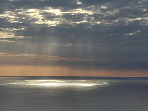 Graues Meer unter dichter Wolkendecke, durch die Lichtstrahlen fallen; im Vordergrund glänzt das Wasser, am Horizont ist ein Schiff zu sehen.