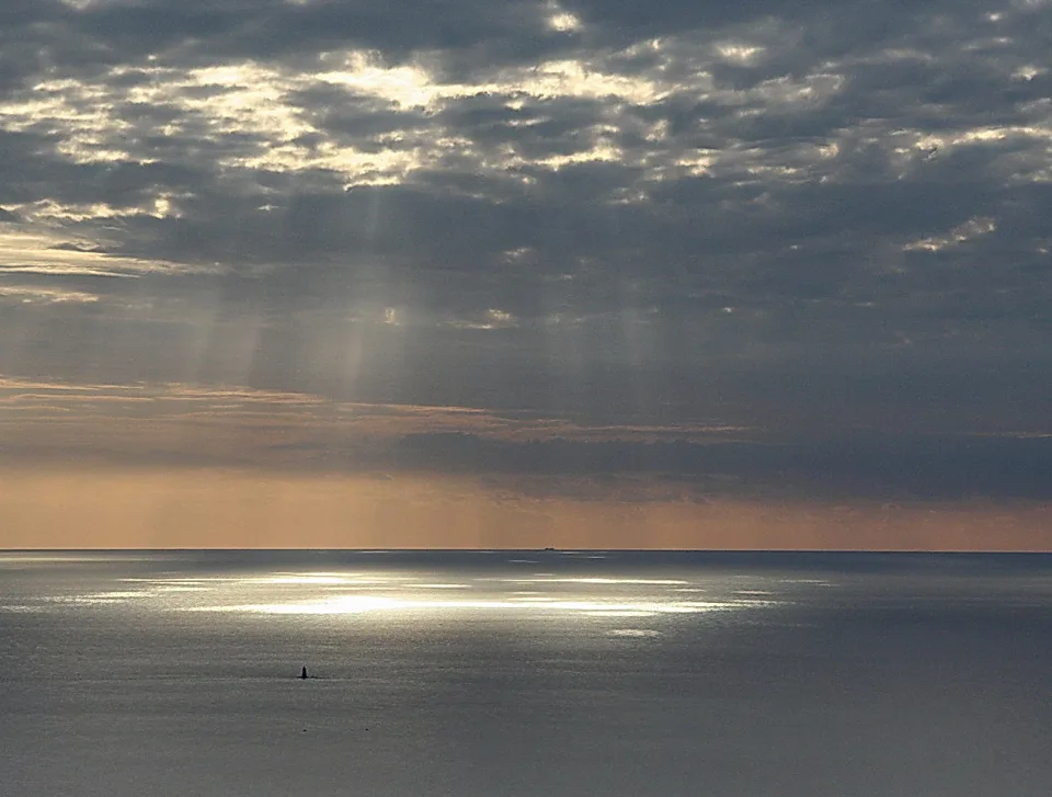 Graues Meer unter dichter Wolkendecke, durch die Lichtstrahlen fallen; im Vordergrund glänzt das Wasser, am Horizont ist ein Schiff zu sehen.