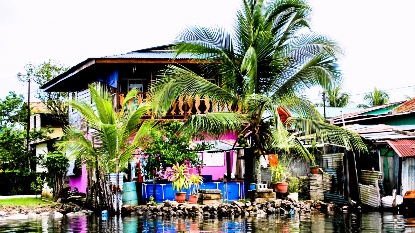 Mehrstöckiges Holzhaus mit pinkfarbener Fassade und Balkon auf einem Pfahl am Ufer, daneben hohe Kokospalmen, Wellblechdächer und ein blauer Wassertank vor einer Steinmauer am Wasser.