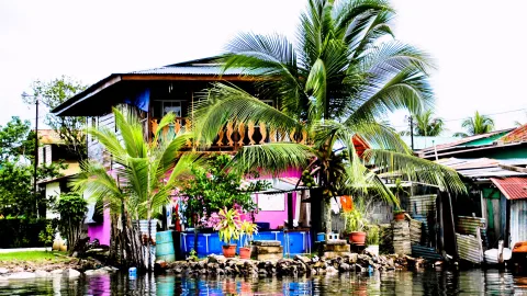 Mehrstöckiges Holzhaus mit pinkfarbener Fassade und Balkon auf einem Pfahl am Ufer, daneben hohe Kokospalmen, Wellblechdächer und ein blauer Wassertank vor einer Steinmauer am Wasser.