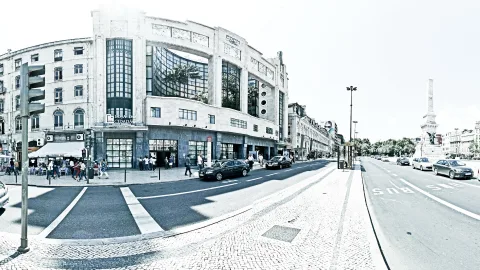 Breite Straßenkreuzung in Lissabon mit einem weiß-grauen Jugendstil-Gebäude mit Schild Loja do Cidadão, Ampel links, Autos auf der Fahrbahn und ein Obelisk rechts im Hintergrund, Bild in Schwarzweiß mit leichtem Blaustich
