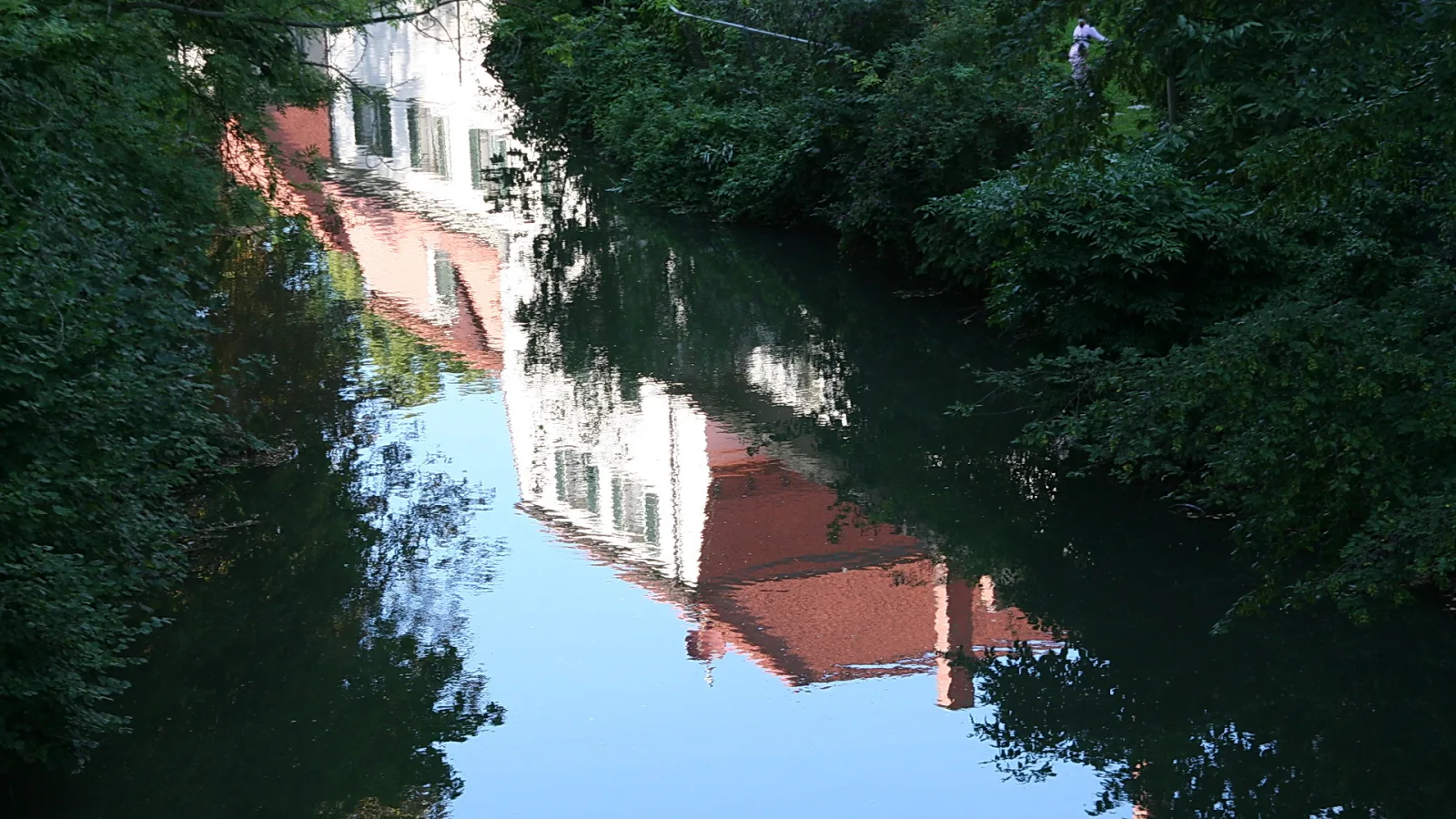 Weiße und rote Häuserfassaden spiegeln sich in einem ruhigen Fluss, der von dichtem grünem Gebüsch gesäumt ist.
