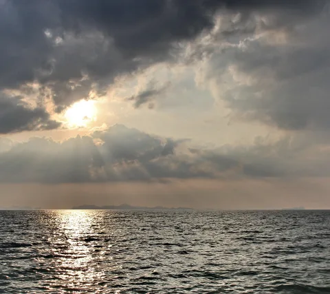 Die Sonne bricht durch dunkle Wolken über einem ruhigen Meer, am Horizont liegen flache Inseln.