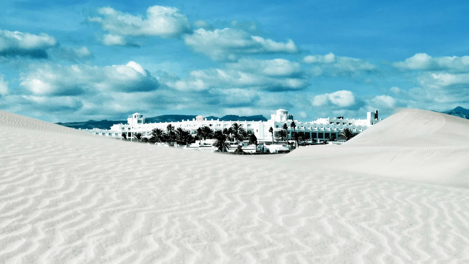 Helle Sanddünen mit Rippenmuster im Vordergrund, dahinter eine lang gezogene weiße Hotelanlage mit Palmen unter blauem Himmel mit Wolken.