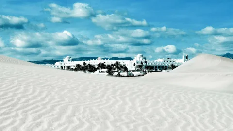 Helle Sanddünen mit Rippenmuster im Vordergrund, dahinter eine lang gezogene weiße Hotelanlage mit Palmen unter blauem Himmel mit Wolken.