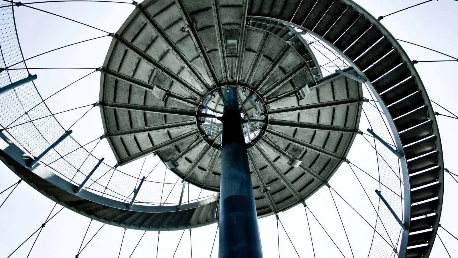 Blick von unten in die Spirale des Killesberg-Aussichtsturms in Stuttgart, mit einem dunklen Mittelpfosten, runden Stahlplattformen, Geländern und einem Netz aus Drahtseilen vor hellem Himmel.