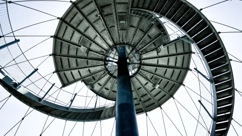 Blick von unten in die Spirale des Killesberg-Aussichtsturms in Stuttgart, mit einem dunklen Mittelpfosten, runden Stahlplattformen, Geländern und einem Netz aus Drahtseilen vor hellem Himmel.