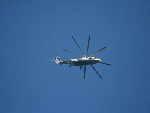Ein weiß-roter Transporthubschrauber mit acht Rotorblättern fliegt von links nach rechts vor einem wolkenlosen blauen Himmel.