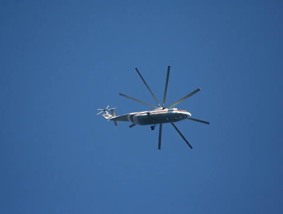 Ein weiß-roter Transporthubschrauber mit acht Rotorblättern fliegt von links nach rechts vor einem wolkenlosen blauen Himmel.
