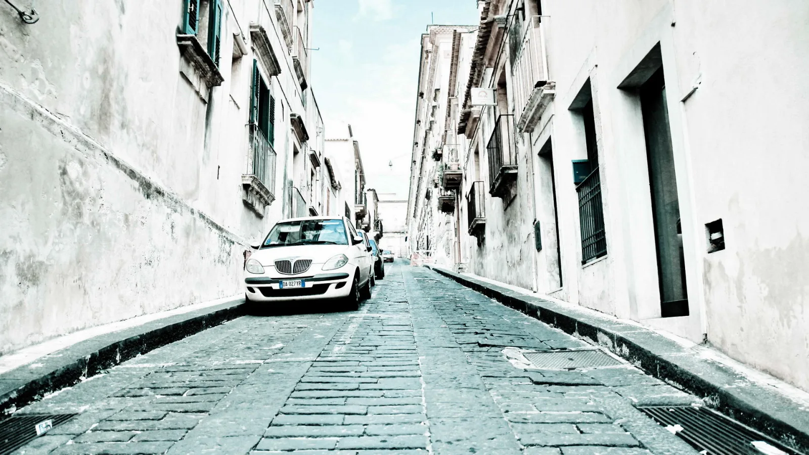 Schmale Kopfsteinpflastergasse zwischen verputzten weißen Altbauten mit Balkonen, links parkt ein weißes Auto, am Ende der Gasse ist heller Himmel zu sehen.