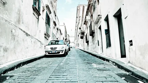 Schmale Kopfsteinpflastergasse zwischen verputzten weißen Altbauten mit Balkonen, links parkt ein weißes Auto, am Ende der Gasse ist heller Himmel zu sehen.