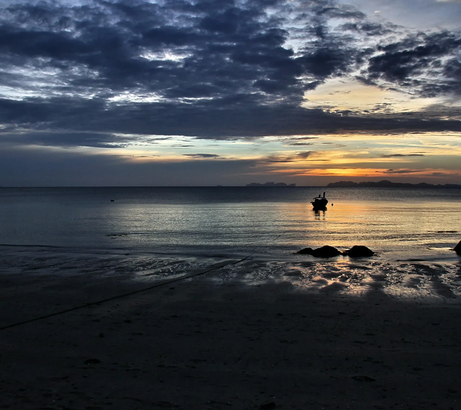 Silhouette eines einzelnen Bootes auf ruhigem Meer bei Sonnenuntergang, davor ein flacher Sandstrand mit dunklen Felsen unter einem bewölkten Himmel.