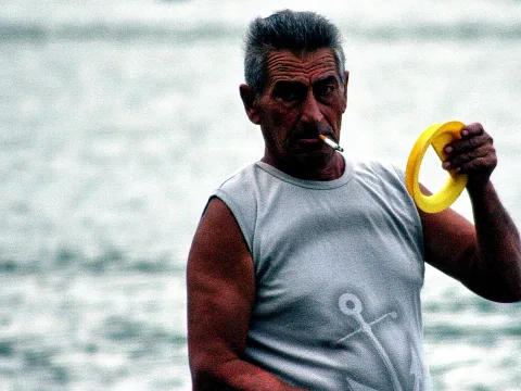 Älterer Mann mit grauen Haaren und Zigarette im Mund hält einen gelben Ring vor einem graugrünen Meer.