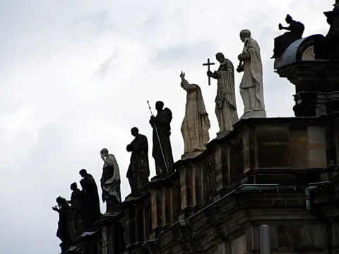 Mehrere steinerne Heiligenfiguren stehen auf einer Balustrade vor bewölktem Himmel, die meisten als dunkle Silhouetten, drei davon hell beleuchtet.