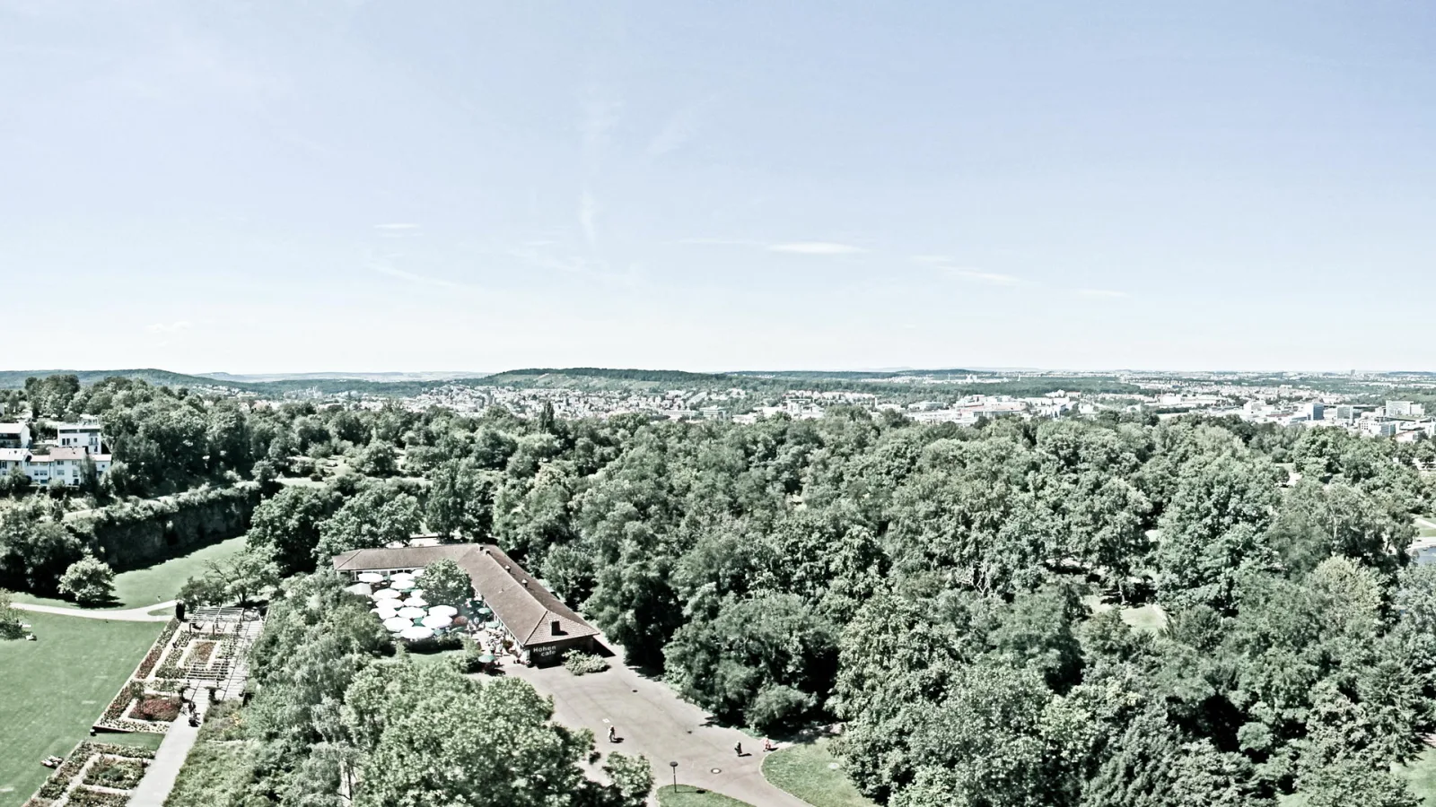 Luftaufnahme eines bewaldeten Parks mit einem Gebäude unter Sonnenschirmen, dahinter eine Stadt unter hellblauem Himmel.