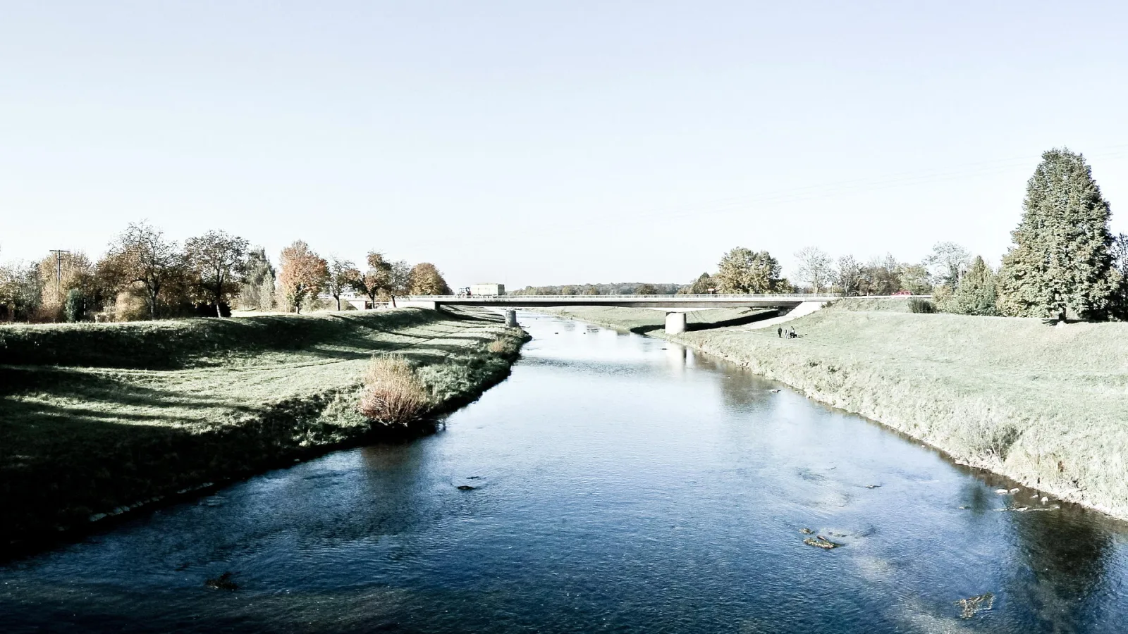 Ein ruhig fließender Fluss führt von vorn nach hinten durch eine flache Landschaft. Eine Betonbrücke überspannt ihn, links und rechts stehen herbstlich gefärbte Bäume unter einem hellen, wolkenlosen Himmel.