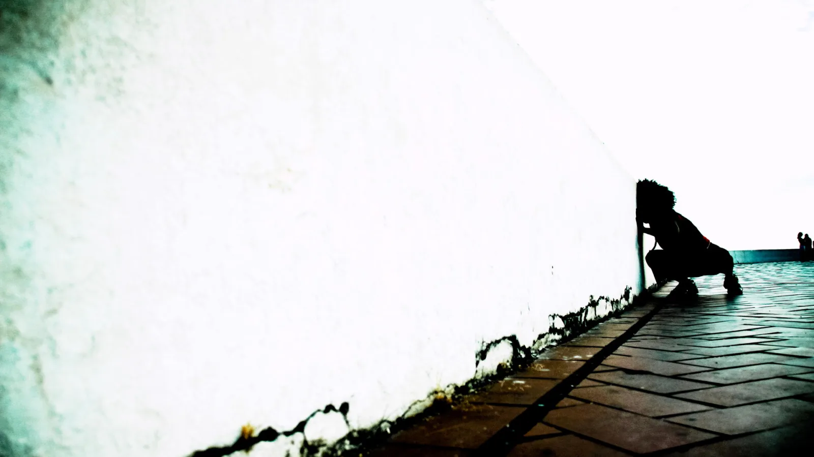 Schwarze Silhouette einer hockenden Person vor einer hellen, fast weißen Wand, rechts ein gepflasterter Weg mit einer weiteren kleinen Figur am Bildrand