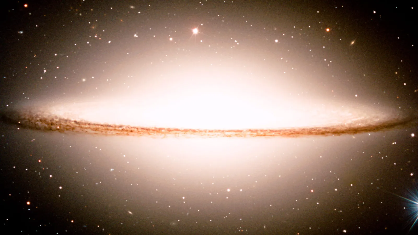Eine Spiralgalaxie in Seitenansicht, als leuchtender Wulst mit dunklem, staubigem Band in der Mitte, umgeben von einem Sternenfeld vor schwarzem Hintergrund.