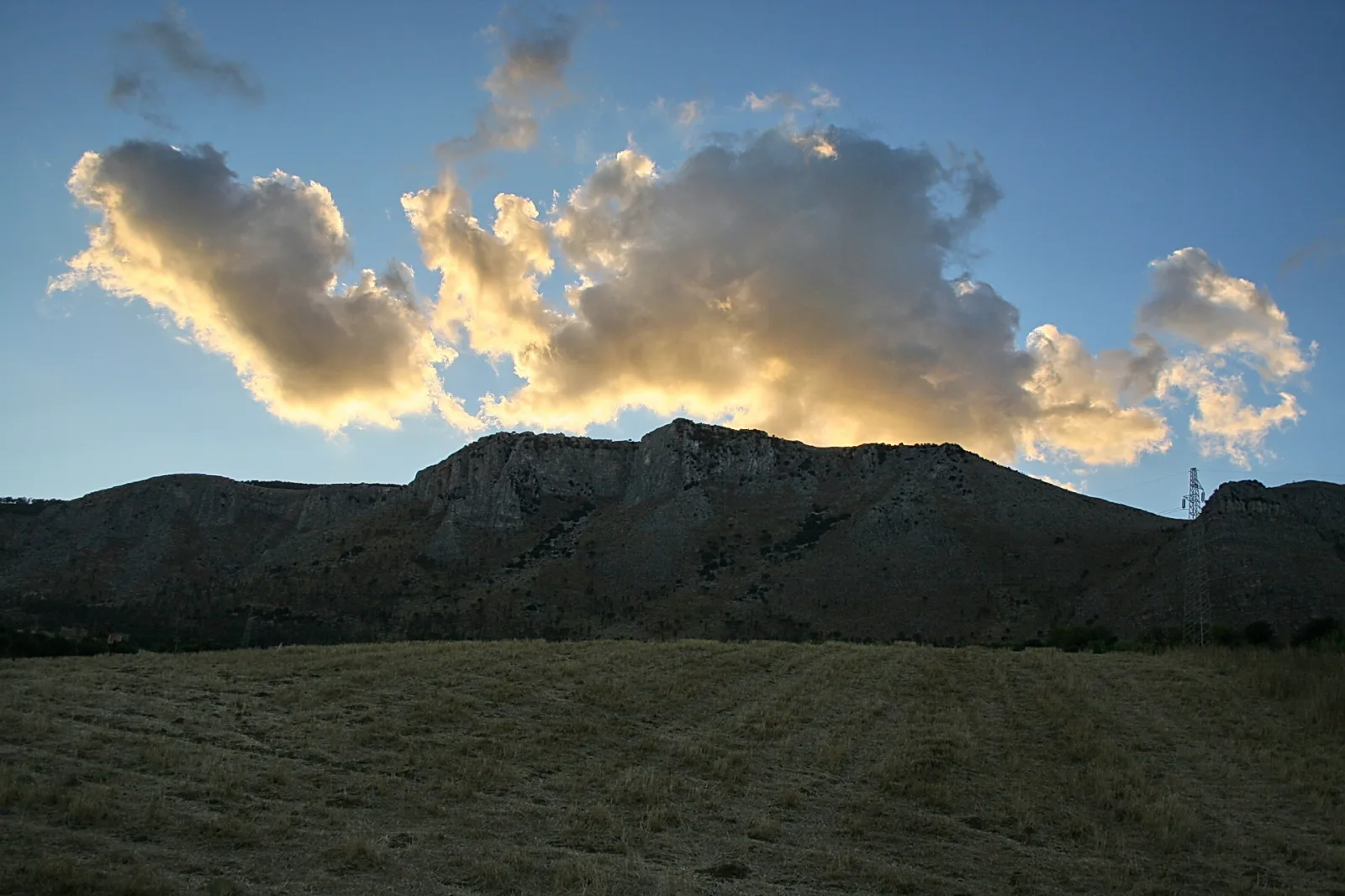 Felsiges Bergmassiv mit hellen, von der Abendsonne angestrahlten Wolken darüber, davor eine abgeerntete, braungrüne Grasebene.