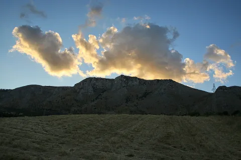 Felsiges Bergmassiv mit hellen, von der Abendsonne angestrahlten Wolken darüber, davor eine abgeerntete, braungrüne Grasebene.